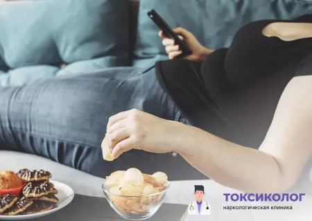 Человек лежит на диване с телефоном и вредной едой, что является показанием для начала программы «Лечение пищевой зависимости»